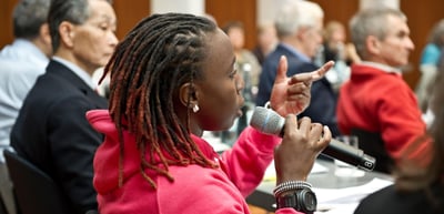 Young woman in a pink jacket speaking into a microphone during a discussion