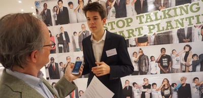 Teenage boy holding a smartphone and a paper in front of a poster with the slogan "Stop talking, start planting"