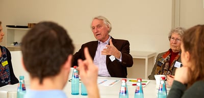 A laureate speaking at a round table while a student raises a hand