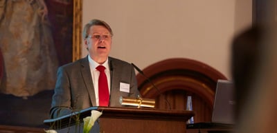 Man in a grey suit and red tie speaking at a lectern
