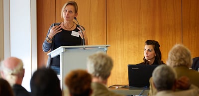 Woman speaking at a lectern, a moderator at a laptop beside her