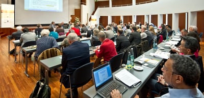 Participants working at long tables with laptops and papers in a large conference hall