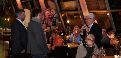 Guests at dinner tables in a restaurant, a man speaking into a microphone
