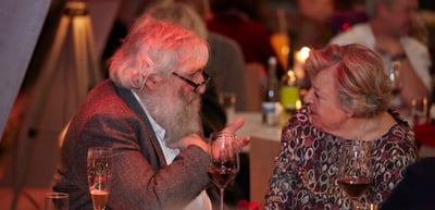 Older man with a white beard and a woman talking over wine at a candlelit table