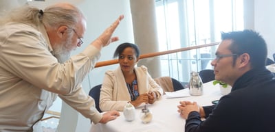 Older man with a white beard leaning over a table to talk with a woman and a man