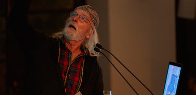 Older man with a white beard and a knitted cap speaking at a lectern, pointing upwards