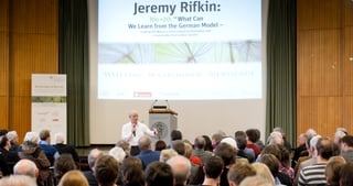 Speaker in front of a full lecture hall, the screen reads "Jeremy Rifkin: Rio+20: What Can We Learn from the German Model, Leading the Way to a Third Industrial Revolution and a Sustainable Post Carbon Society"