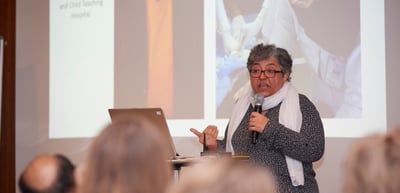 Woman with glasses and a white scarf speaking into a microphone next to a projected slide
