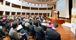 Full audience in a round lecture hall listening to a speaker at a lectern