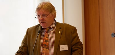 Man with glasses in a brown jacket speaking at a lectern