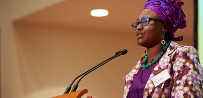 Woman in a colourful dress and head wrap speaking at a lectern