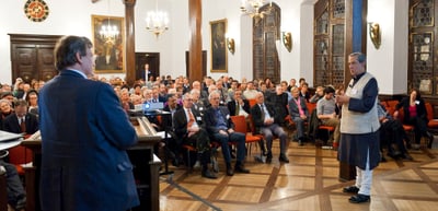 Speaker at a lectern addressing a seated audience in a historic hall with wood panelling and chandeliers