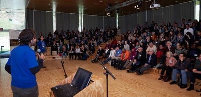 Young man in a blue shirt speaking in front of a full audience in a hall with wooden steps