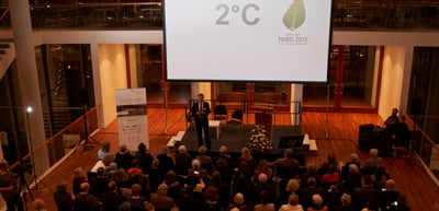 Audience in the foyer of the Konzerthaus Freiburg in front of a stage, a slide shows 2°C and Paris 2015