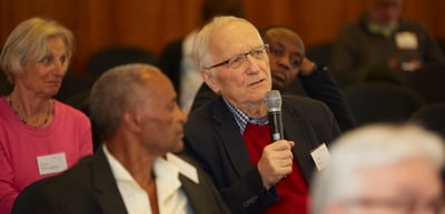 Older man with glasses speaking into a microphone from his seat in the audience