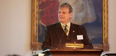 Man with glasses speaking at a lectern in front of a large painting