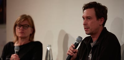 Man speaking into a microphone next to a woman with glasses during a podium talk