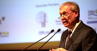 Man with glasses speaking at a lectern in front of a projected slide with partner logos