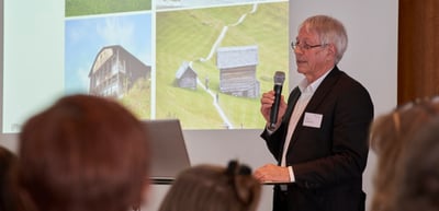 A laureate speaking into a microphone in front of a slide with photos of wooden huts in a mountain landscape