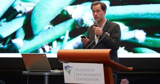 Man speaking into a microphone at a lectern of the European Environment Foundation in front of a large projected photo of plants