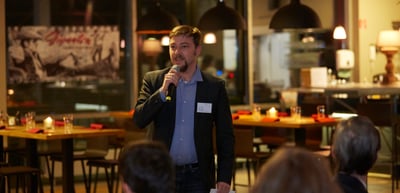 Man speaking into a microphone in a restaurant with candles on the tables