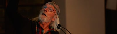 Older man with a white beard and a knitted cap speaking at a microphone and looking upwards
