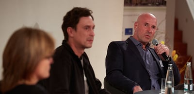 Three people in a podium talk, a bald man speaking into a microphone