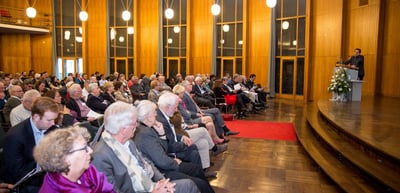 Audience seated in rows in a hall with wood panelling and tall windows, a speaker at a lectern on the stage