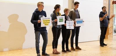 Students on stage holding signs with the names and titles of the impulse lectures by Oana Elena Stürz, Rupert Ebner, Deborah Klein Walker and Stephan Schindele