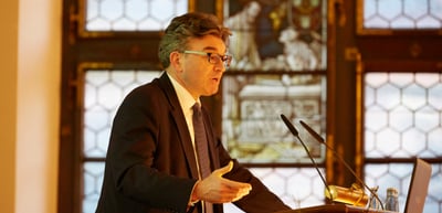 Man with glasses speaking at a lectern in front of stained glass windows