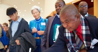 Man in a checked scarf bending over a table to write, other participants standing behind him