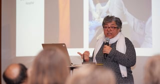 Woman with glasses and a white scarf speaking into a microphone next to a projected slide