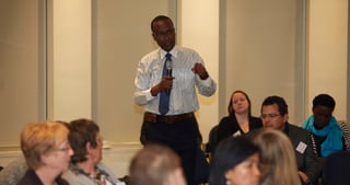 Man in a shirt and tie speaking to a seated audience in a hall with columns