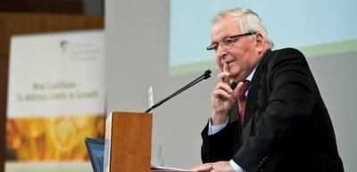 Older man with glasses at a lectern, resting his chin on his hand, in front of a blurred banner reading "New Coalitions, To Address Limits to Growth"
