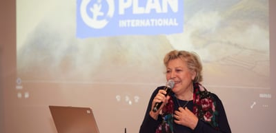 Woman speaking into a microphone in front of a slide with the logo of Plan International