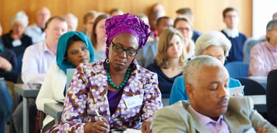 Woman in a colourful dress and head wrap taking notes in a full audience