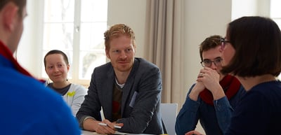A man in a grey jacket listening at a round table with students