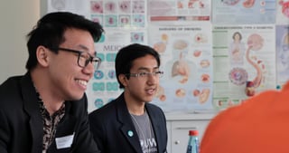 Two smiling young men at a round table in a classroom with posters on the wall
