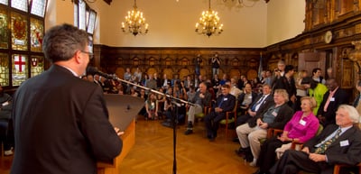 Speaker at a microphone stand addressing the audience in a historic hall with chandeliers