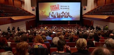 Full concert hall in front of a cinema screen reading "Power to Change, Welcome to the premiere!"