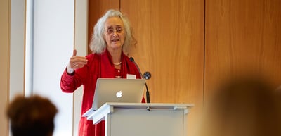 Woman with white curly hair in a red jacket speaking at a lectern with a laptop