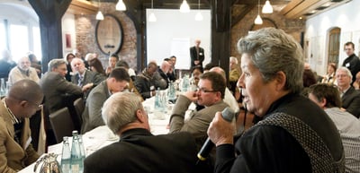 Discussion round at long tables in a hall with wooden beams, a woman speaking into a microphone