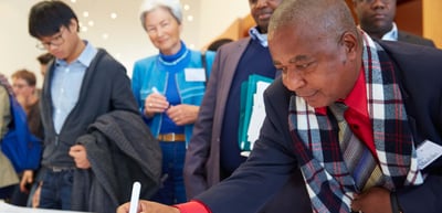 Man in a checked scarf bending over a table to write, other participants standing behind him