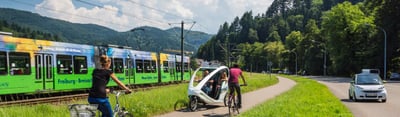 Tram, cyclists, a bicycle taxi and a car on a road in Freiburg with wooded hills