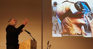 Michael Wadleigh speaking at a lectern next to a projected photo of a person with a camera