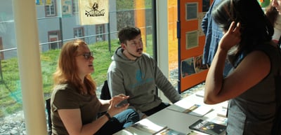 Young woman and young man sitting at an exhibition table talking with a visitor