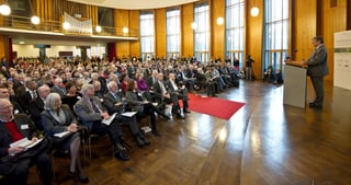 Speaker at a lectern in a wood-panelled hall in front of a large audience