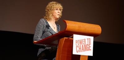Woman with curly blond hair speaking at a lectern with the sign "Power to Change"