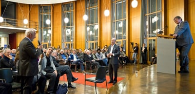 Speaker at a lectern answering a question from a man with a microphone in a hall with tall windows