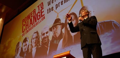 Man speaking into a microphone in front of the screen with the poster of "Power to Change, Die Energierebellion"